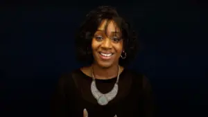 Smiling woman with curly hair wearing a black top and statement necklace against a dark background.