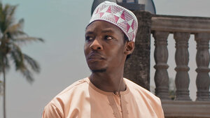 Man in traditional attire stands against a tropical backdrop with palm trees and stone architecture in bright sunlight.