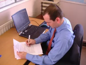 Businessman reviewing documents while sitting at a desk with a laptop and stationery in an office setting.