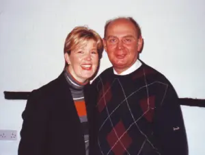 Smiling couple posing together in a cozy indoor setting, dressed in casual sweaters against a neutral-colored wall.