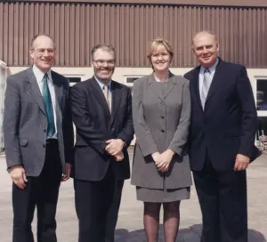 Four professionals standing together outdoors, smiling, in front of a building, dressed in smart business attire.