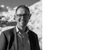 Smiling man in glasses wearing a blazer, standing against a snowy mountain backdrop. Bright and cheerful atmosphere.