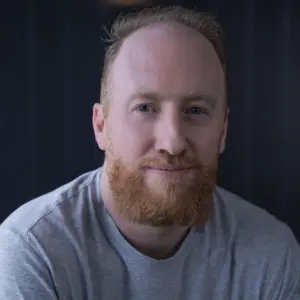 Smiling man with a beard wearing a gray shirt, sitting against a dark background with soft lighting.