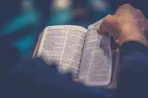 Person holding an open book with visible text, symbolizing study or contemplation in a softly lit environment.