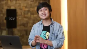 Young person in a denim jacket smiling and gesturing, standing by a laptop in a modern, well-lit workspace.