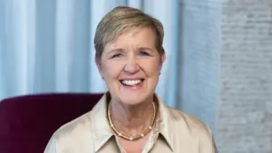 Smiling woman with short blonde hair wearing a light blouse, seated against a light-colored background, exuding warmth.