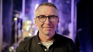 Smiling man in glasses with a blurred band backdrop, conveying a welcoming and engaging atmosphere.