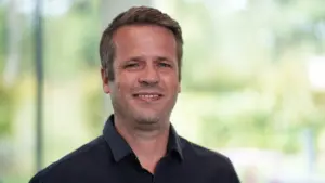 Smiling man in a dark shirt standing indoors with a blurred green background, exuding confidence and approachability.