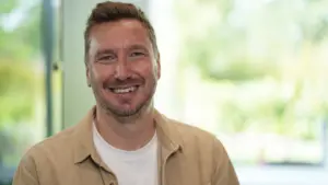 Smiling man in a casual beige shirt, set against a bright, natural background, conveying warmth and approachability.