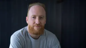 Man with a red beard and short hair smiling against a dark background, wearing a gray t-shirt.