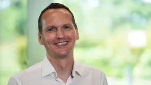 Smiling man in a light shirt stands against a vibrant green blurred background, embodying positivity and confidence.