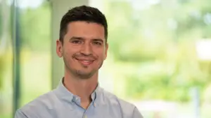 Smiling young man in a light blue shirt with a blurred outdoor background, conveying approachability and professionalism.