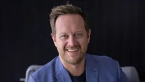 Smiling man in a blue blazer poses confidently against a dark background, showcasing a warm and approachable demeanor.