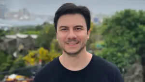 Smiling man with dark hair standing outdoors, greenery in the background, cityscape visible in the distance.