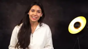 Smiling woman in a white zip-up sweatshirt standing next to a stylish yellow lamp against a dark background.