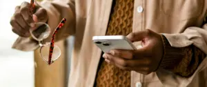 Person holding a smartphone and glasses, wearing a patterned shirt and a light jacket in a modern setting.