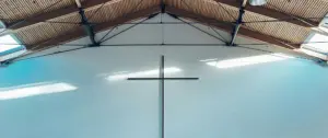 Interior view of a church featuring a large wooden cross against a bright white wall and a rustic ceiling.