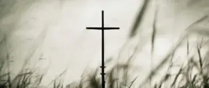 Silhouette of a cross against a dramatic gray sky, surrounded by soft grass and blurred foreground elements.