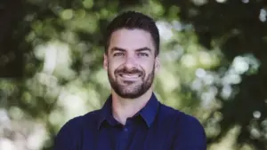 Smiling man with beard in a blue shirt stands outdoors against a blurred green background.