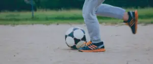 Child playing soccer on a sandy field, dribbling a black and white ball with orange-accented sneakers.