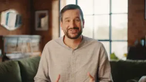 Smiling man in casual shirt gesturing warmly, seated in a cozy living room with large windows and greenery.