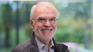 Smiling elderly man with glasses and a beard in a blurred outdoor setting, wearing a light jacket.