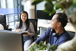 Two colleagues collaborating in a modern office, one taking notes and the other engaging in conversation beside a plant.