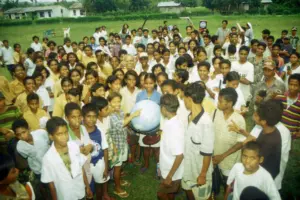 Crowd of diverse children and adults gathered outdoors, holding a globe amid a vibrant green landscape.