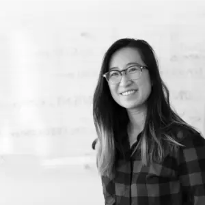 Smiling woman in glasses wearing a plaid shirt, standing in front of a whiteboard with handwritten notes.