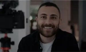 Smiling man with a beard in front of a camera, showcasing a bright, inviting atmosphere in an indoor setting.