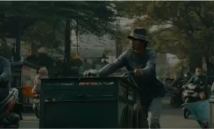 Man in a hat pushing a cart through a bustling street, surrounded by traffic and greenery, capturing urban life dynamics.