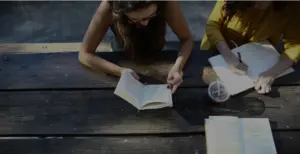 Two women studying together at a wooden table, one reading a book while the other takes notes and enjoys a drink.